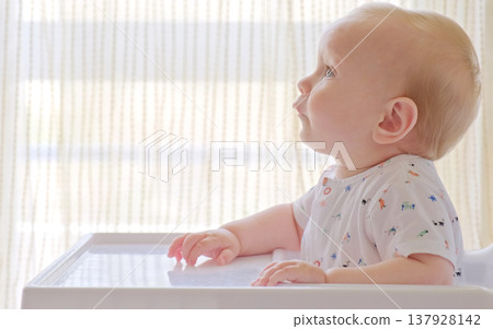 Cute baby is sitting on a high chair waiting for a meal. Beautiful white-haired toddler is sitting in a high children's chair at home. 137928142