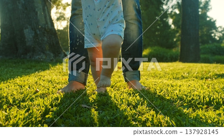 Walking children's bare feet on a green lawn close-up. Child learns to take the first steps on the grass. Baby learns to walk with the help of his mother on a green grass in the park. Walking children's bare feet on a green lawn close-up. Child learns to take the first steps on the grass. Baby learns to walk with the help of his mother on a green grass in the park. 137928145