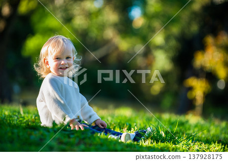 Happy cute toddler enjoying a summers day in a park while sitting on the grass. Smiling adorable boy sitting on the grass in a park on a sunny day. Cheerful small child alone on the lawn and looking 137928175