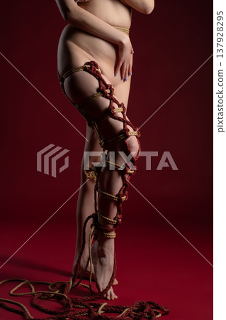 Intricate shibari rope bondage adorns a woman's leg and torso in a dramatic studio setting with a red background. 137928295