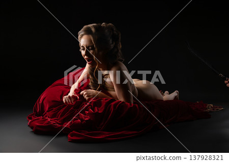 Passionate woman in artistic rope bondage lying on rich red velvet in a dark studio setting. 137928321