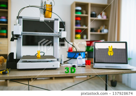 A 3D printer is actively creating a small yellow bunny figure on its platform, with a laptop displaying the design nearby A 3D printer is actively creating a small yellow bunny figure on its platform, with a laptop displaying the design nearby 137928674