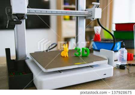 A modern 3D printer is shown with a small yellow bunny figure and green 3D letters on its build plate, ready for creation 137928675