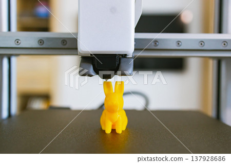A close-up shot of a 3D printer nozzle depositing material to create a small yellow bunny figurine on its build plate A close-up shot of a 3D printer nozzle depositing material to create a small yellow bunny figurine on its build plate 137928686