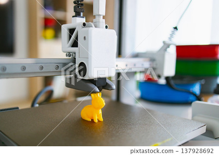 A modern 3D printer is in the process of creating a small, yellow, bunny-shaped toy on its build plate 137928692