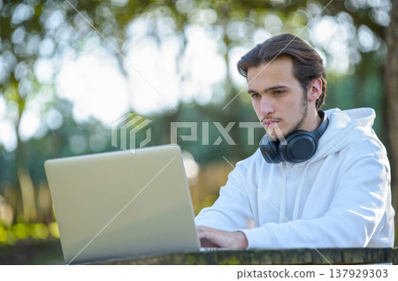 Student is working at a laptop in the park. Young guy is typing on a computer keyboard in a park sitting at a table. 137929303