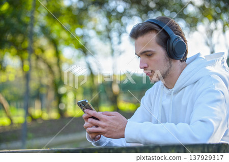 Portrait of young man in headphones with a phone in his hands outdoors. Young guy looks at screen of a cell phone in the park. Handsome man with smartphone listen to the music with headphones Portrait of young man in headphones with a phone in his hands outdoors. Young guy looks at screen of a cell phone in the park. Handsome man with smartphone listen to the music with headphones 137929317