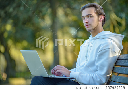 Portrait of thinking student at the park with laptop on his legs looks away. Portrait of thinking student at the park with laptop on his legs looks away. 137929329