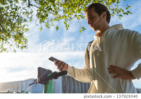 Student scanning the electric scooter's QR-code by phone. Man points a mobile-phone camera to scan a scooter's printed code. Using an app to rent an environmentally friendly green electric scooter. 137929340
