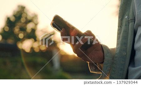 Hands using mobile over sunset. Close-up of men's hands with a mobile in their hands typing sms. Person is typing a text message on a cell phone phone - close-up. People and communication technology. 137929344
