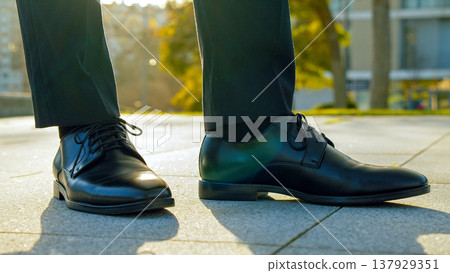 Close-up view to the businessman in a black new classic shoes outdoors. Legs of businessman in fashionable glossy shoes on the sidewalk. Business concept. Stylish men wears. Low angle. Close-up view to the businessman in a black new classic shoes outdoors. Legs of businessman in fashionable glossy shoes on the sidewalk. Business concept. Stylish men wears. Low angle. 137929351