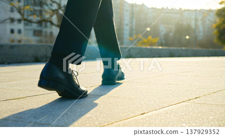 Legs of a businessman in fashionable shoes walking outdoors. Business concept. Close-up view to the businessman in a black new shoes walks on the street. Stylish men wears. Low angle. Rear view. Legs of a businessman in fashionable shoes walking outdoors. Business concept. Close-up view to the businessman in a black new shoes walks on the street. Stylish men wears. Low angle. Rear view. 137929352