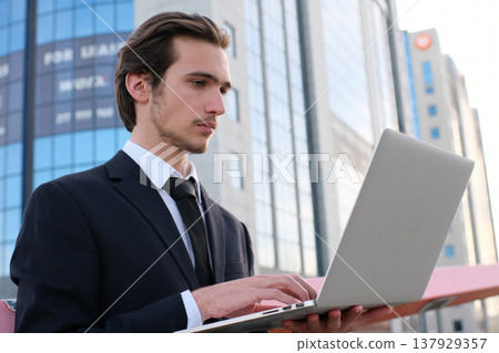 Confident businessman near a business center with a computer. A young businessman in a suit is looking at a laptop. A young businessman near a modern tall glass building with a camputer. 137929357