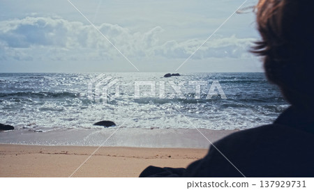 Young man stands on the empty sandy beach on the summer day and contemplating ocean water surface. Human stands on beach on a sunny day and looking to distance. Back view of Man on the ocean shore 137929731
