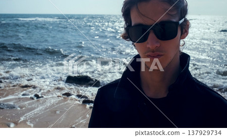 Young man with blown brown hair against the ocean on summer sunny day. Attractive young man looks at the camera on the beach. Handsome guy in black sunglasses poses outdoors on vacation. Young man with blown brown hair against the ocean on summer sunny day. Attractive young man looks at the camera on the beach. Handsome guy in black sunglasses poses outdoors on vacation. 137929734