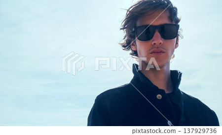 Handsome guy with long hair poses against sky with sun rays. Stylish man in black jacket looks at the camera, low angle shot. Young man with blown brown hair against the blue sky on summer sunny day. Handsome guy with long hair poses against sky with sun rays. Stylish man in black jacket looks at the camera, low angle shot. Young man with blown brown hair against the blue sky on summer sunny day. 137929736