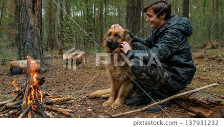 Young man with big dog sits at the fire in the forest. Cheerful guy is stroking his dog, outdoor. Happy caucasian teenager with his German shepherd by the fire in nature, in autumn. 137931232