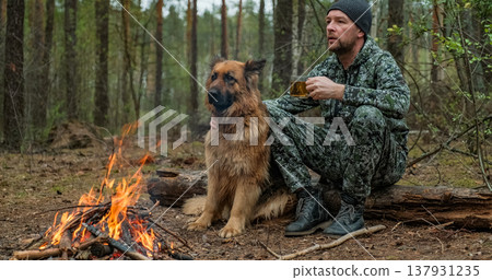 Adult Man is relaxing at the fire with his dog in the nature. Alone traveler is resting with his German shepherd by the forest fire and drinking tea. Man strokes a dog at the fire in the forest. 137931235