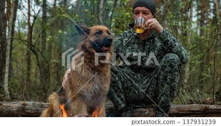 Adult Man is relaxing at the fire with his dog in the nature. Alone traveler is resting with his German shepherd by the forest fire and drinking tea. Man strokes a dog at the fire in the forest. 137931239