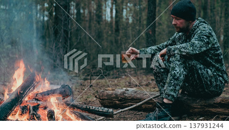 Man with cup of tea the fire in the forest. Human looks at the bonfire and drinks tea on the nature in the summer. Alone traveler sits and rests by the fire with a cup of coffee at dusk. Man with cup of tea the fire in the forest. Human looks at the bonfire and drinks tea on the nature in the summer. Alone traveler sits and rests by the fire with a cup of coffee at dusk. 137931244