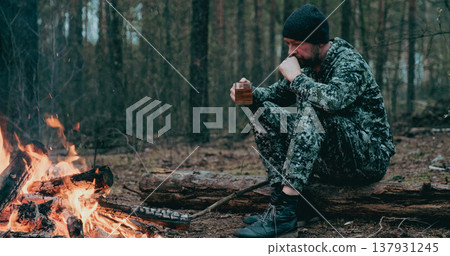 Man with cup of tea the fire in the forest. Human looks at the bonfire and drinks tea on the nature in the summer. Alone traveler sits and rests by the fire with a cup of coffee at dusk. Man with cup of tea the fire in the forest. Human looks at the bonfire and drinks tea on the nature in the summer. Alone traveler sits and rests by the fire with a cup of coffee at dusk. 137931245