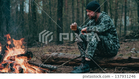 Man with cup of tea the fire in the forest. Human looks at the bonfire and drinks tea on the nature in the summer. Alone traveler sits and rests by the fire with a cup of coffee at dusk. 137931246