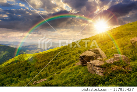 sunset over beautiful summer landscape in mountains. grassy hill with huge stones under the cloudy sky and rainbow. wonderful nature background. picture of hillside meadow near the mountain peak 137933427