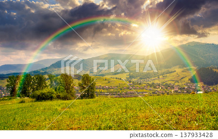 sunset over summer landscape with grassy pasture in mountains. beautiful countryside imagery with settlement in the distant valley under cloudy sky and rainbow. spectacular rural scenery 137933428