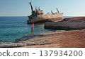 Woman stands on rocky Mediterranean shore near Paphos, Cyprus, gazing at the rusty EDRO III shipwreck grounded off the coast under a clear blue sky, serene travel scene 137934200