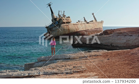 Woman stands on rocky Mediterranean shore near Paphos, Cyprus, gazing at the rusty EDRO III shipwreck grounded off the coast under a clear blue sky, serene travel scene 137934200
