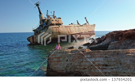 Tourist sitting on a rocky cliff watching the grounded rusty cargo ship EDRO III against the clear blue sky and turquoise Mediterranean Sea near Paphos, Cyprus Tourist sitting on a rocky cliff watching the grounded rusty cargo ship EDRO III against the clear blue sky and turquoise Mediterranean Sea near Paphos, Cyprus 137934201