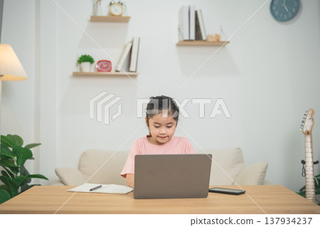 Young girl engaged in online learning at home with a laptop and notebook on a wooden table in a bright, cozy living room environment 137934237