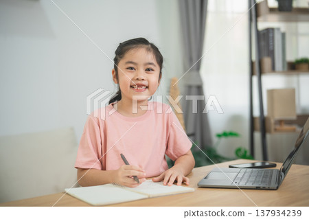 Cheerful young girl engaging in creative learning at home, writing in notebook while sitting at desk with modern laptop and cozy decor in background 137934239