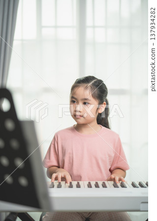 Young girl playing keyboard at home, focused expression, practicing music, indoor setting with bright light, learning and growth concept, educational activity 137934282