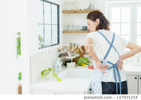 一位中年家庭主婦弓著背坐在廚房裡，摀著背。 137935254