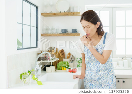 一位中年家庭主婦弓著身子坐在廚房裡，摀著肩膀。 137935256