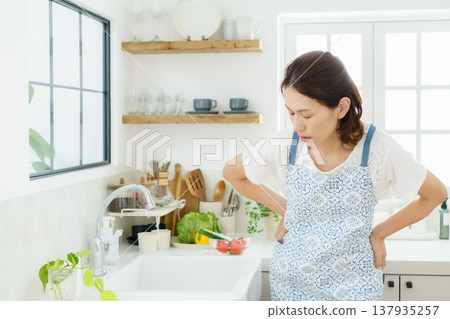 A middle-aged housewife is hunched over in the kitchen, clutching her back. A middle-aged housewife is hunched over in the kitchen, clutching her back. 137935257