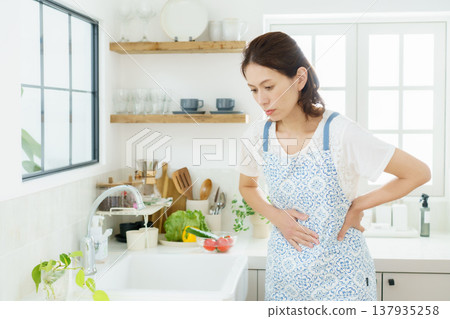 A middle-aged housewife is hunched over in the kitchen, clutching her stomach. A middle-aged housewife is hunched over in the kitchen, clutching her stomach. 137935258