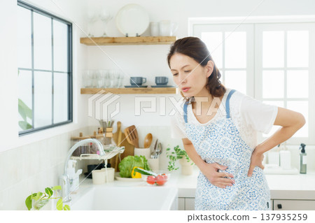 A middle-aged housewife is hunched over in the kitchen, clutching her stomach. A middle-aged housewife is hunched over in the kitchen, clutching her stomach. 137935259