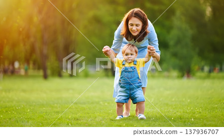 Mother Helping Child Take First Steps in Sunny Park Setting 137936707