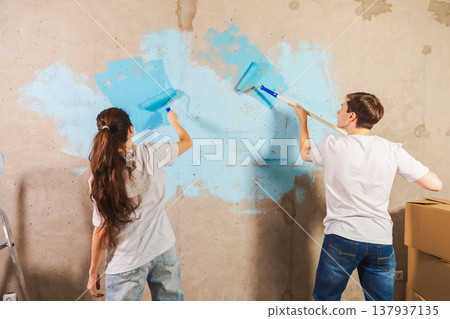 Couple in new home during repair works painting wall together. Happy family holding paint roller painting wall with blue color paint in new house. Home renovation DIY renew home concept 137937135