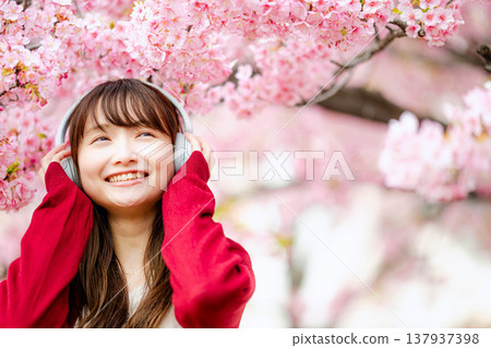 一位年輕女子在櫻花樹下戴著耳機聽音樂。 一位年輕女子在櫻花樹下戴著耳機聽音樂。 137937398