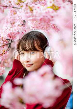 一位年輕女子在櫻花樹下戴著耳機聽音樂。 一位年輕女子在櫻花樹下戴著耳機聽音樂。 137937401