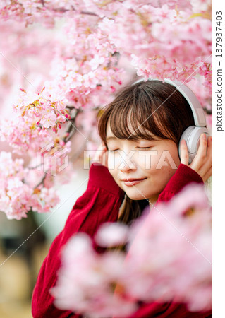 一位年輕女子在櫻花樹下戴著耳機聽音樂。 一位年輕女子在櫻花樹下戴著耳機聽音樂。 137937403