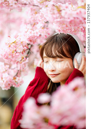 一位年輕女子在櫻花樹下戴著耳機聽音樂。 一位年輕女子在櫻花樹下戴著耳機聽音樂。 137937404
