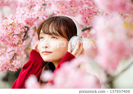 一位年輕女子在櫻花樹下戴著耳機聽音樂。 一位年輕女子在櫻花樹下戴著耳機聽音樂。 137937409