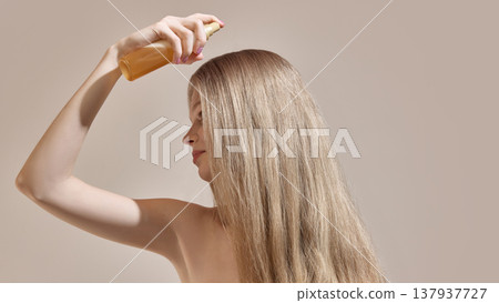 Woman applying spray product to long straight hair in minimal studio environment. Woman applying spray product to long straight hair in minimal studio environment. 137937727