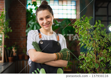 Running of own business. Young woman florist wearing apron in botanical store with green plants. Happy small business owner working at flower shop standing surrounded by plants. Small business Running of own business. Young woman florist wearing apron in botanical store with green plants. Happy small business owner working at flower shop standing surrounded by plants. Small business 137938116