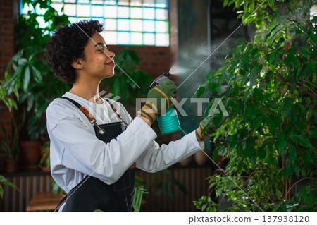 Running of own business. African woman florist with spray bottle watering fertilizing plants in botanical store, Small business owner working at flower shop smiling surrounded by plants Small business 137938120