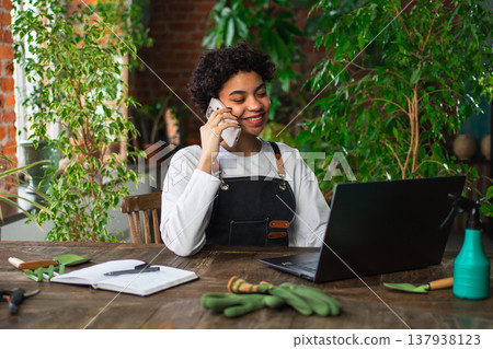 African woman florist gardener working in botanical store using laptop order plants by phone communicating with customer. Female small business owner working at flower shop. Running of own business African woman florist gardener working in botanical store using laptop order plants by phone communicating with customer. Female small business owner working at flower shop. Running of own business 137938123